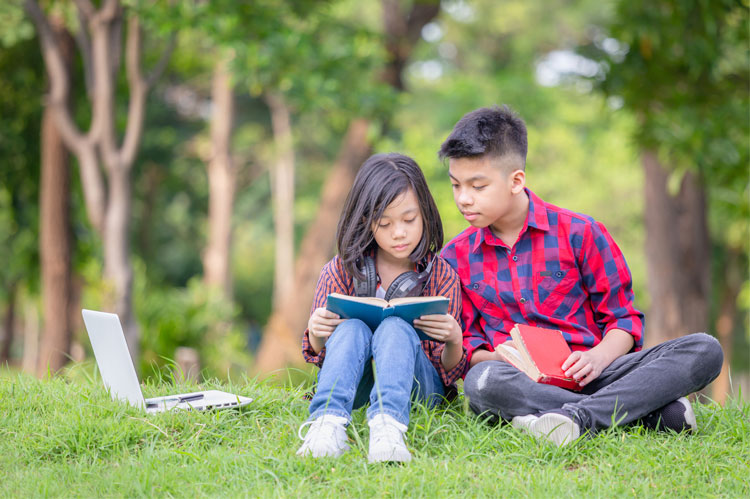 two siblings sit on grass and read book together