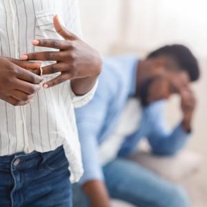person removes wedding ring from finger with person seated in background