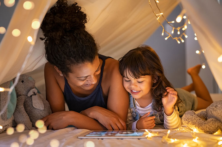 child looks at tablet computer with parent under tent with fairy lights