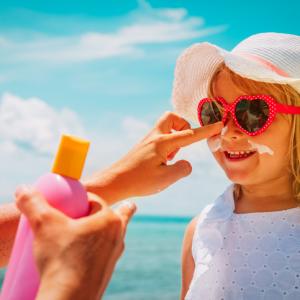 on holiday abroad, mother puts suncream on daughter's face at the seaside. girl wears red sunglasses and white hat