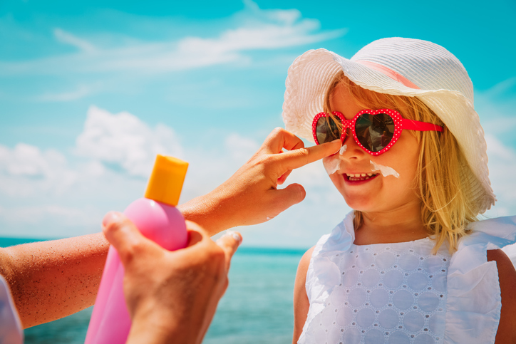 on holiday abroad, mother puts suncream on daughter's face at the seaside. girl wears red sunglasses and white hat