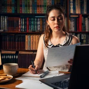 business woman working from home, looking at piece of paper (concept, looking at a cookie complaint) with a laptop and papers on a desk, with bookcase in background