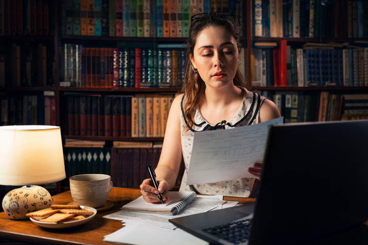 business woman working from home, looking at piece of paper (concept, looking at a cookie complaint) with a laptop and papers on a desk, with bookcase in background