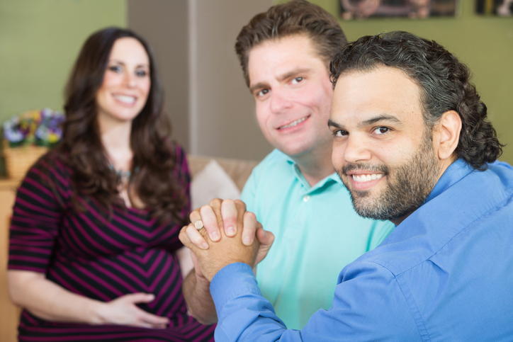 gay couple holding hands, smiling. Pregant woman in the background smiling. Surrogacy concept.