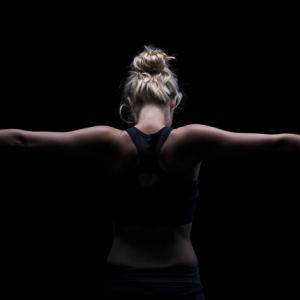 view from the back of a blonde woman with her hair up and arms out, surrounded by darkness. fitness style image. concept: could look like britney spears and represent conservatorship