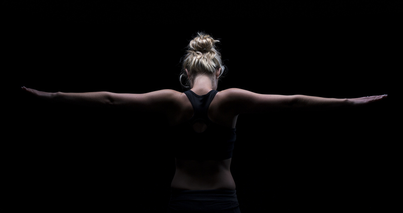 view from the back of a blonde woman with her hair up and arms out, surrounded by darkness. fitness style image. concept: could look like britney spears and represent conservatorship