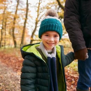 child smiling at camera, outside in autumn woodland, holding hand of adult whose face is cropped out. concept for children contact