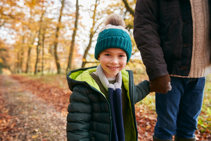 child smiling at camera, outside in autumn woodland, holding hand of adult whose face is cropped out. concept for children contact