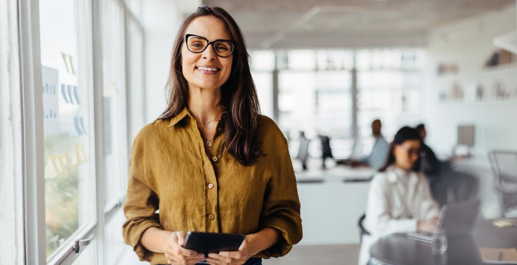 Business owner smiling at the camera in her office while employees work in background