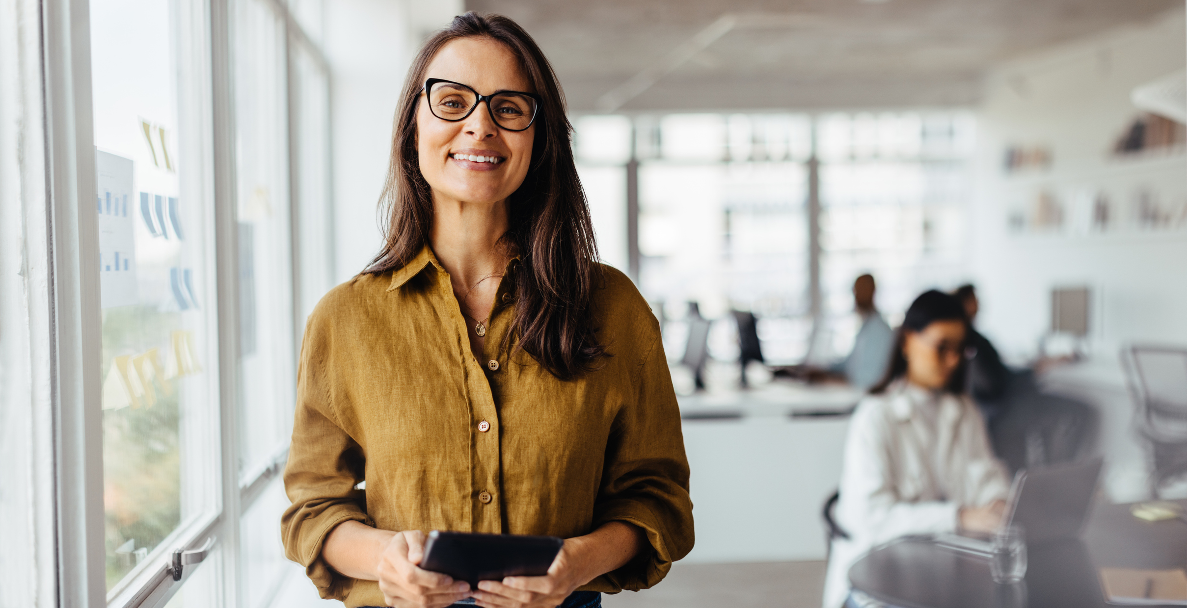Business owner smiling at the camera in her office while employees work in background