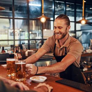 Charming young bartender in apron serving beer and smiling while working in the pub
